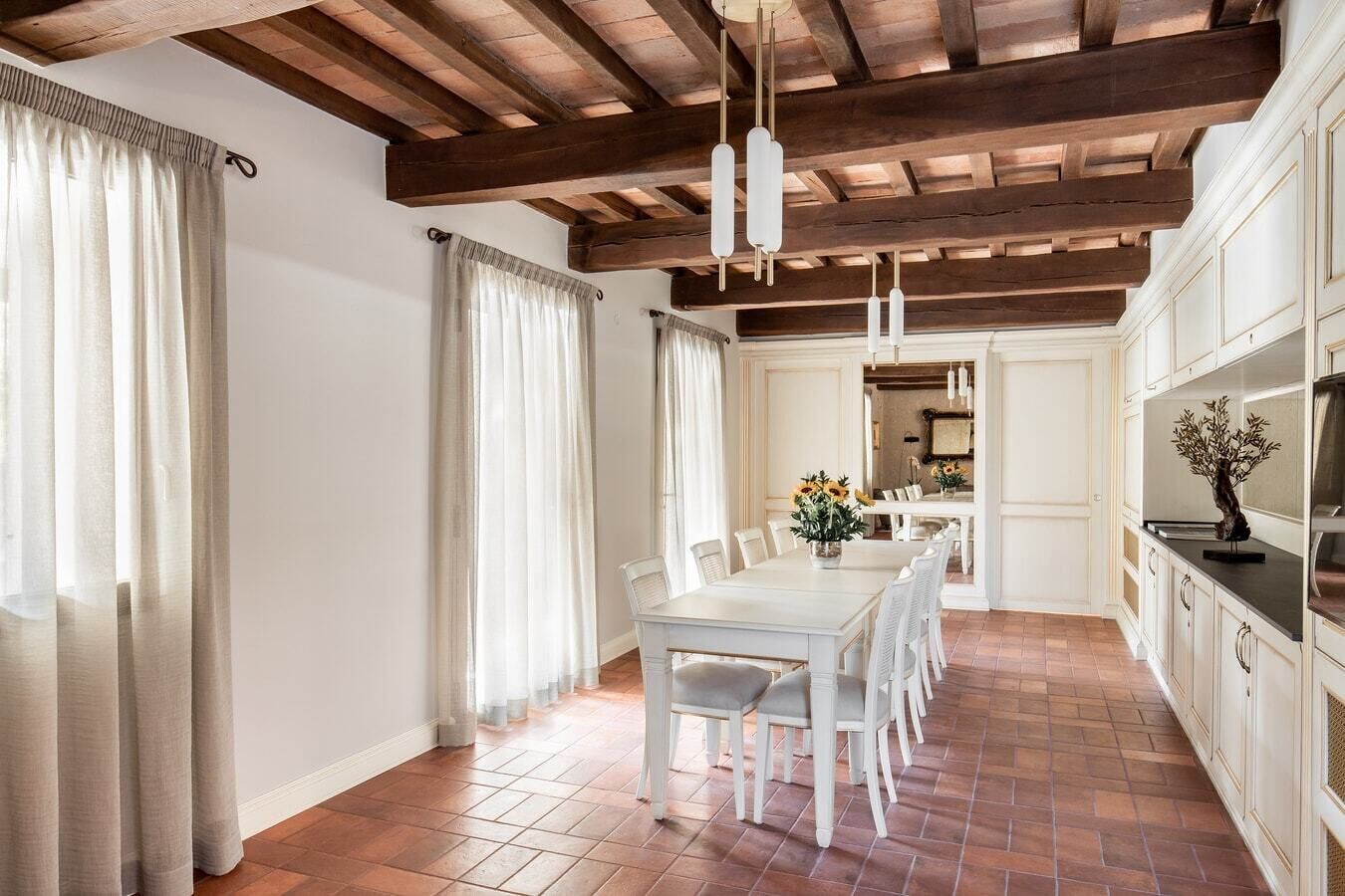 A dining area with a white table and chairs, wooden ceiling beams, and a view into a kitchen area.