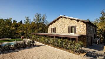 A stone house with a covered outdoor seating area, a swimming pool, and a gravel driveway.