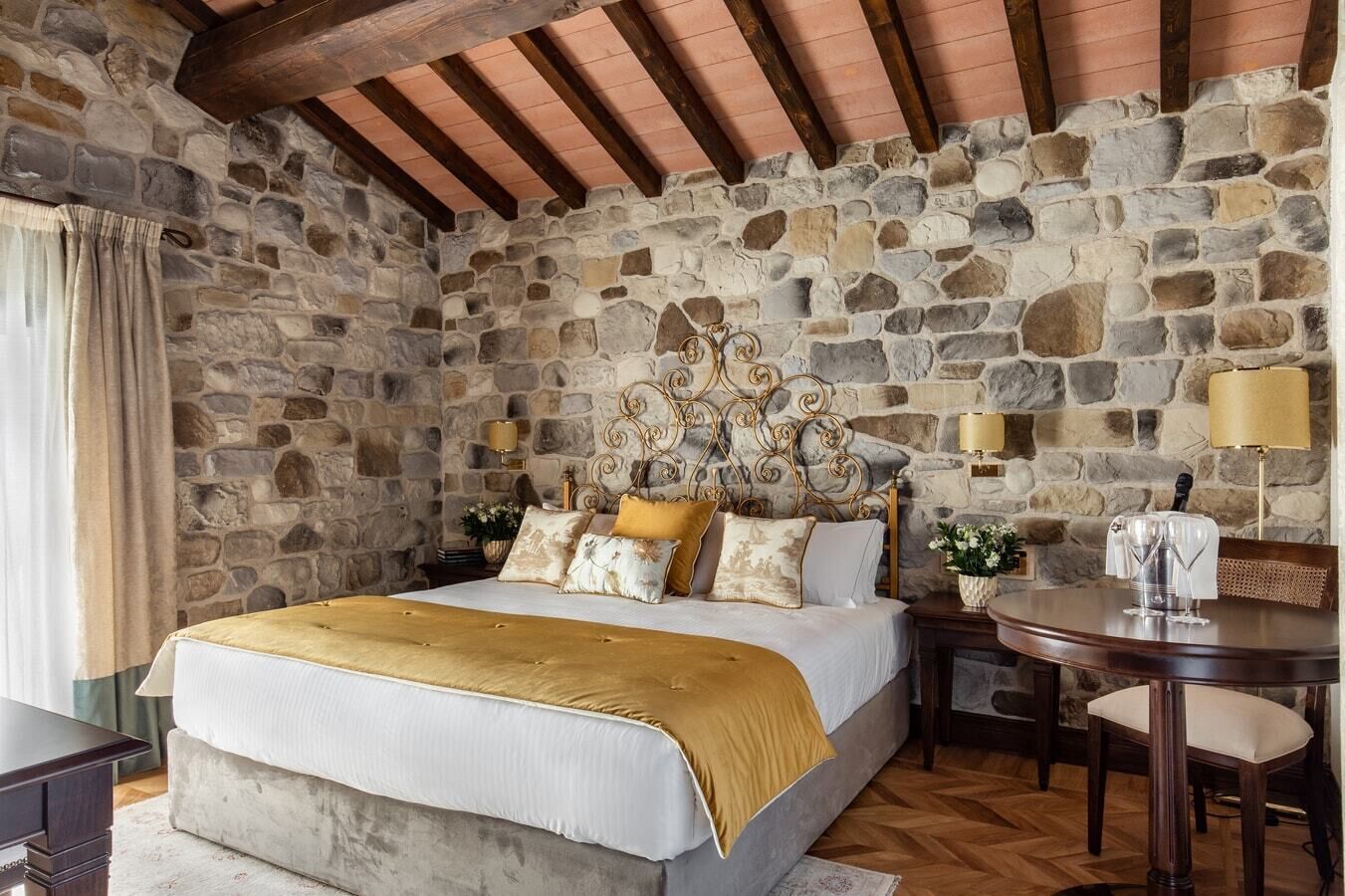 A bedroom with a stone wall, a bed with a gold headboard, a wooden table, and a lamp.