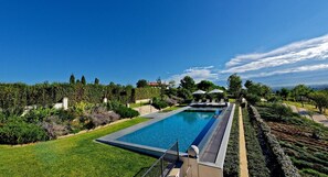 Une piscine rectangulaire à l’eau d’un bleu limpide, entourée de pelouse et de haies parfaitement entretenues, d’où l’on aperçoit un bâtiment et des arbres au loin, sous un ciel dégagé.