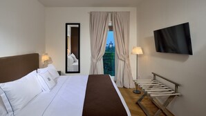Quarto de hotel com cama, televisão, espelho, janela com cortinas e um banco.