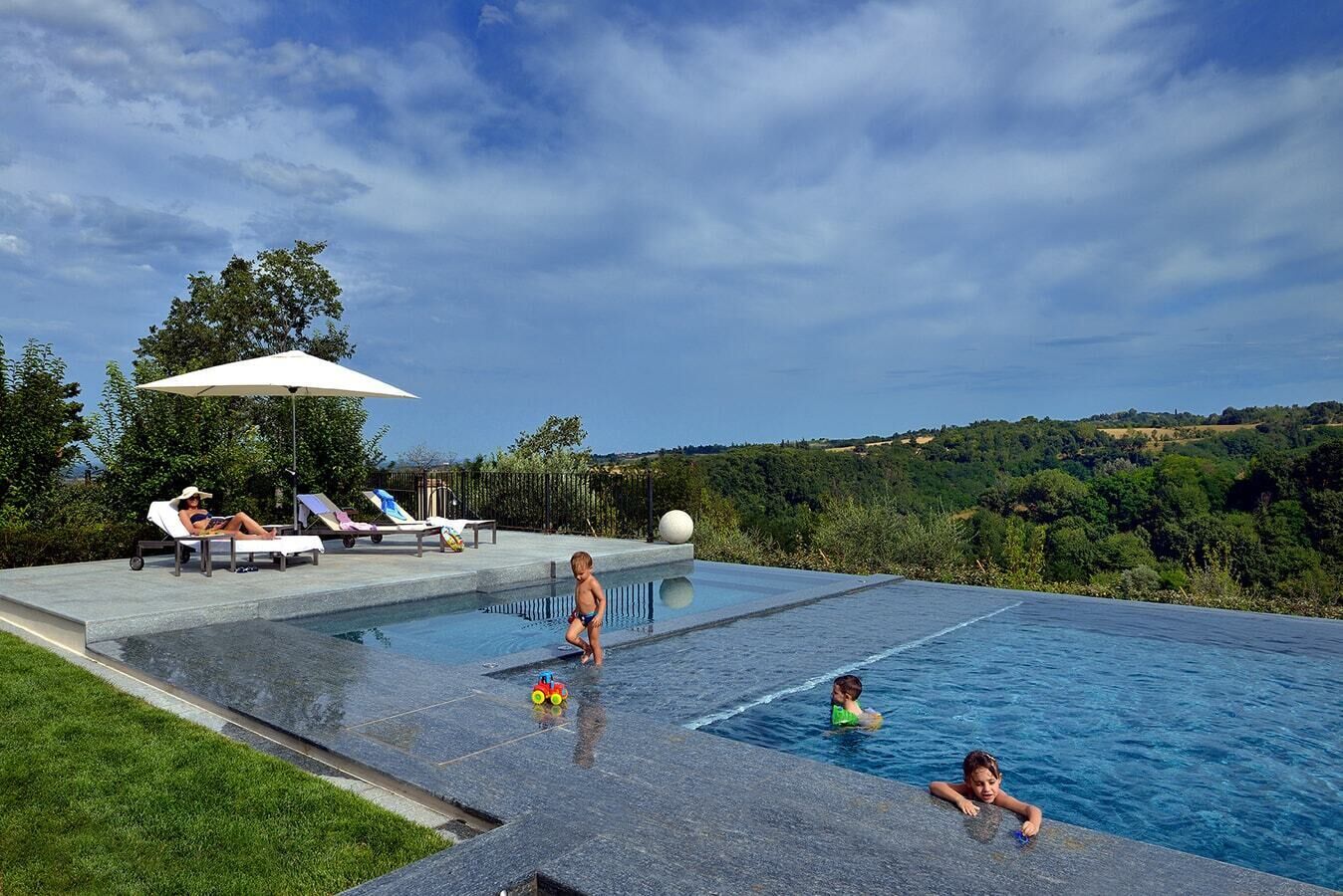 A swimming pool with a view of a hilly landscape, lounge chairs, and a person relaxing.