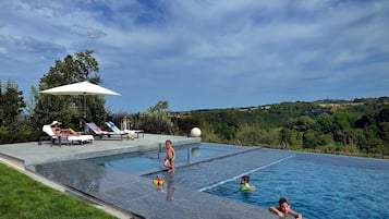 En swimmingpool med udsigt til et bakket landskab, liggestole og en person, der slapper af.
