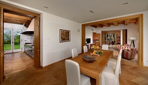 Un vaste espace de salle à manger avec une table en bois et des chaises blanches, une cuisine visible à travers une porte ouverte, et une vue sur l’extérieur.