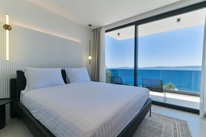 Ein modernes Schlafzimmer mit einem großen Bett, Meerblick und einem Balkon mit Sitzgelegenheiten.