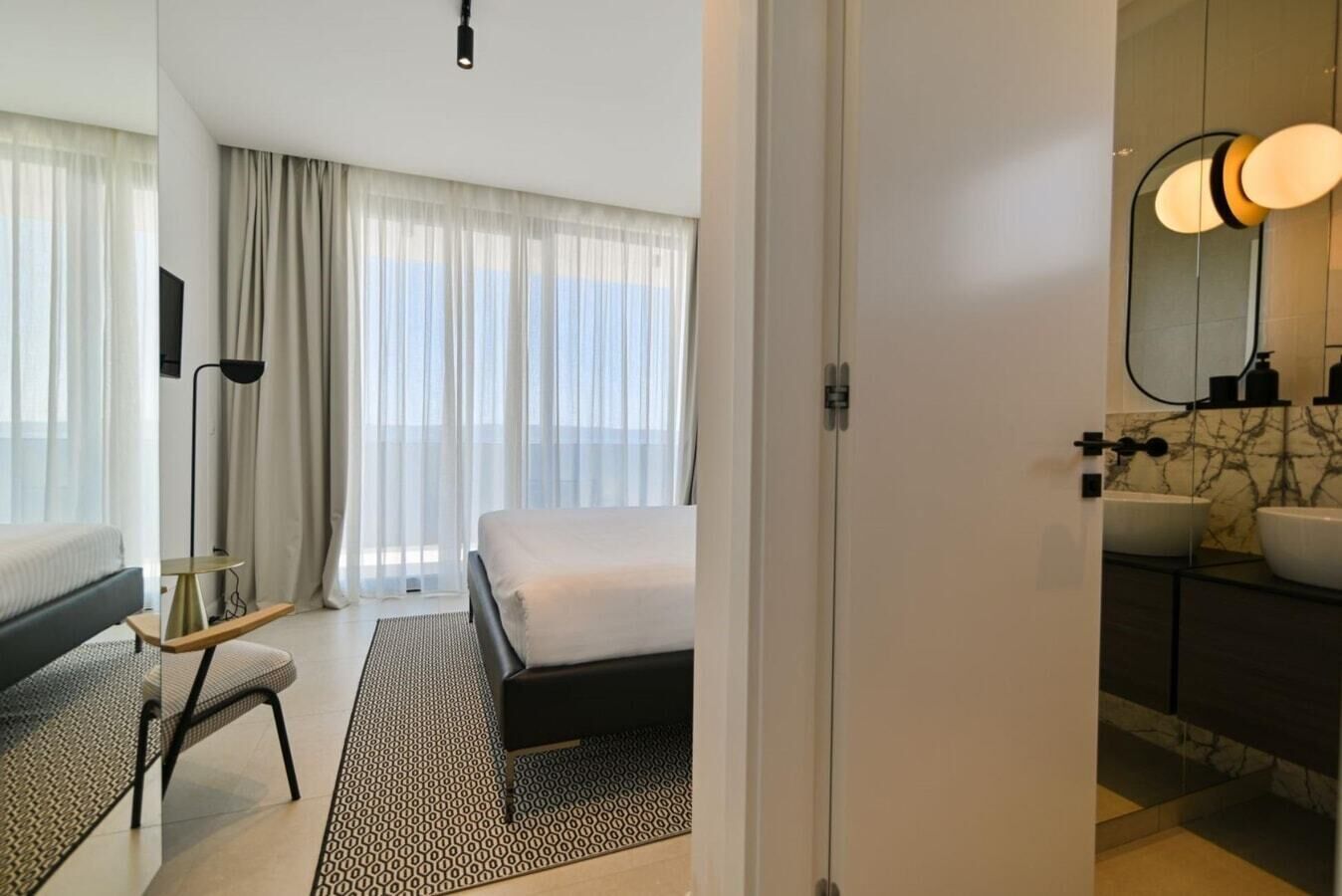 A hotel room with a bed, bedside table, lamp, and a view of the outdoors through sheer curtains.
