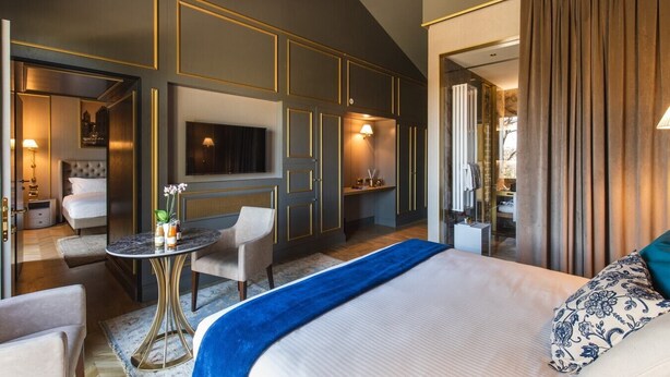 Ein modernes Hotelzimmer mit einem großen Bett, einer Sitzecke mit Tisch, einem an der Wand befestigten Fernseher und einem durch eine Glastür einsehbaren Badezimmer.