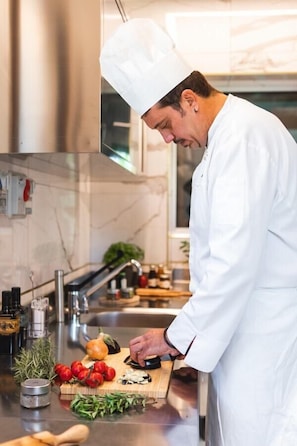 A chef in a white uniform preparing food in a modern kitchen with stainless steel appliances and a marble backsplash.