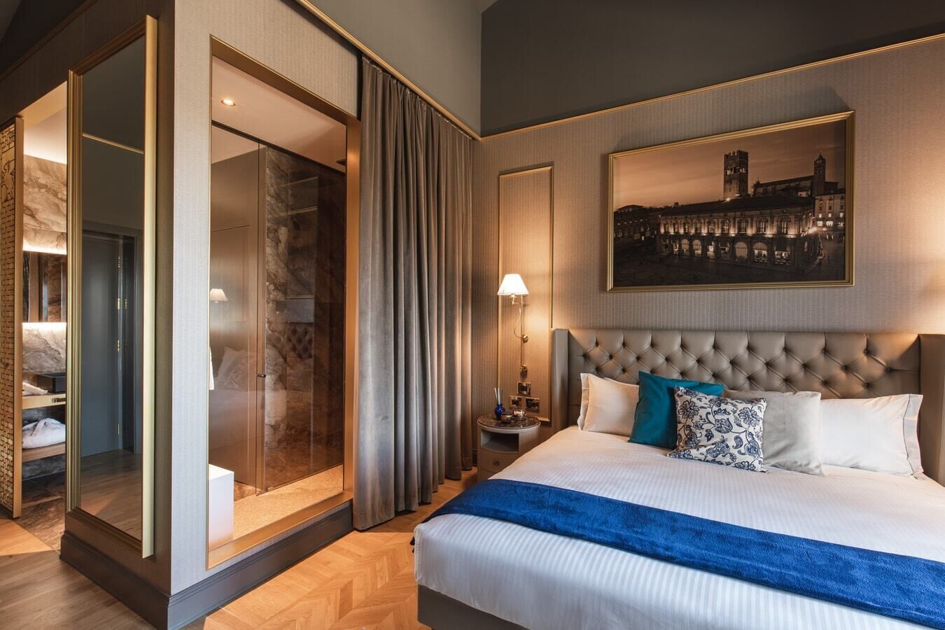 A hotel room with a large bed, a nightstand, a lamp, a wardrobe with a mirror, and a cityscape painting on the wall.