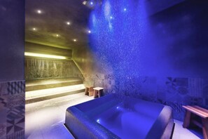 Ein Wellnessraum mit einer großen Badewanne, einem kleinen Holzschemel und einer Wand mit gemusterten Fliesen.