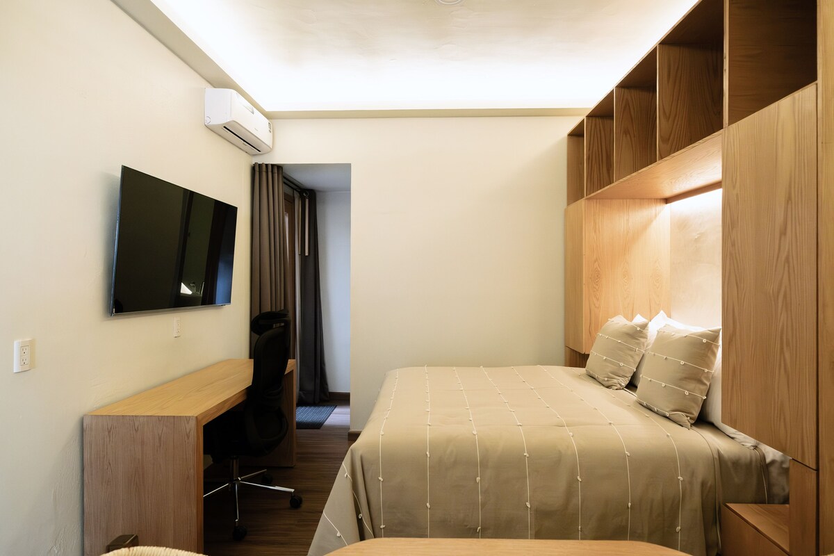 A hotel room with a bed, a desk with a chair, a television, and an air conditioning unit.