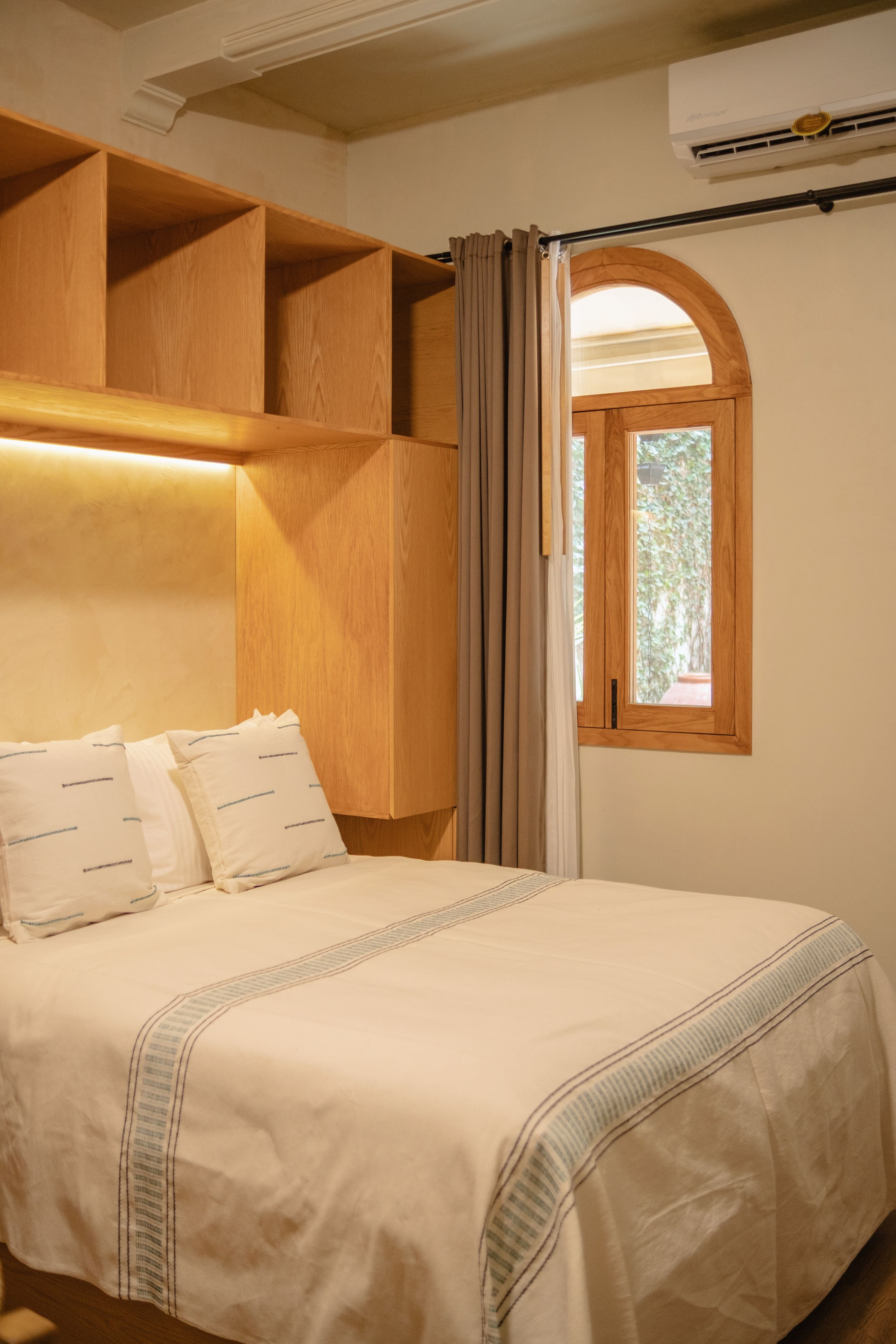 Un dormitorio con cama, cabecera de madera, ventana con cortinas y un equipo de aire acondicionado.
