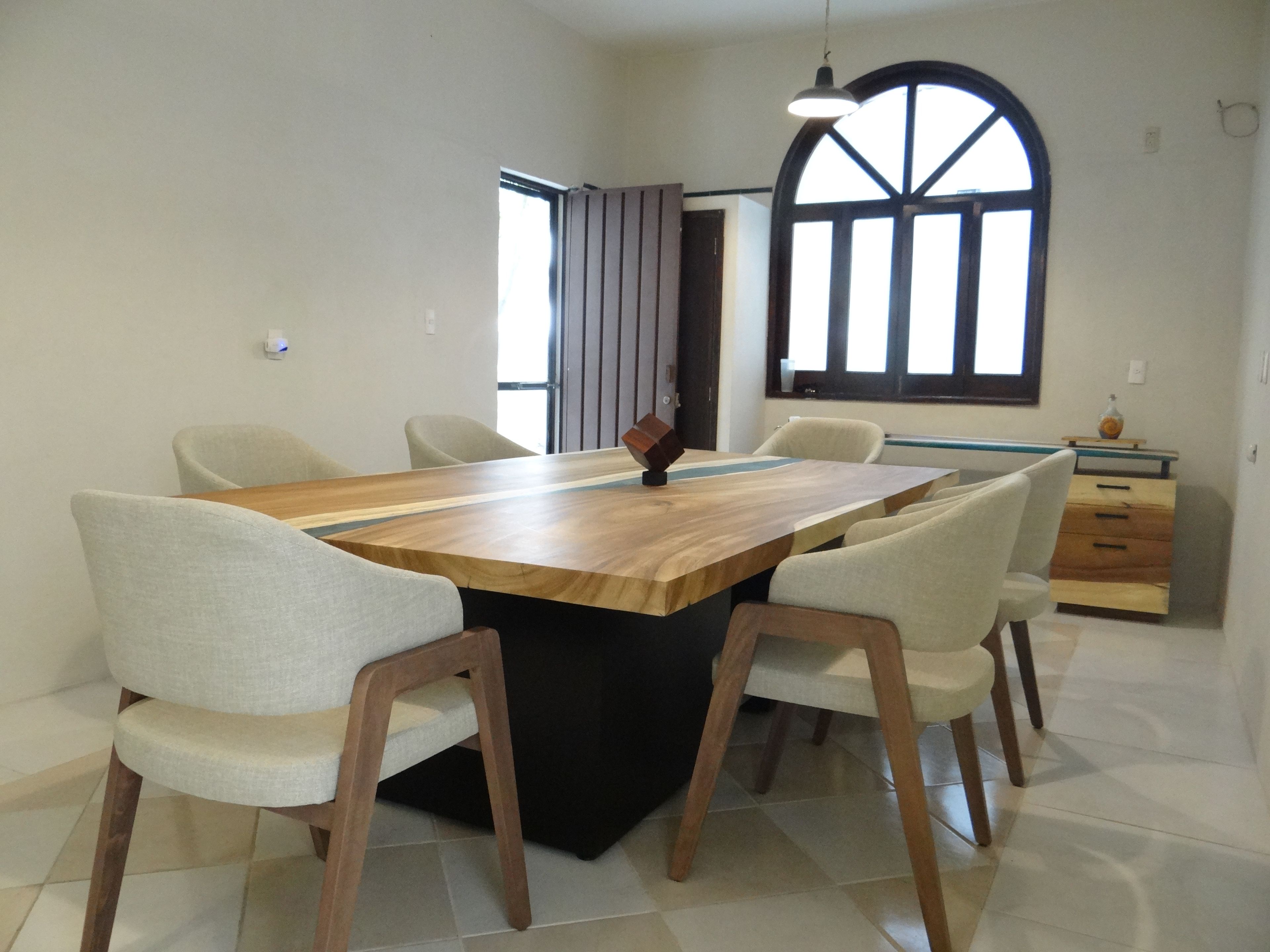 Un comedor con una mesa de madera rectangular rodeada por seis sillas, un ventanal con cortinas y un mueble bajo con una botella encima.