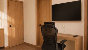 Una habitación con un escritorio de madera, una silla de oficina negra, un televisor de pantalla plana montado en la pared y una puerta de madera cerrada.