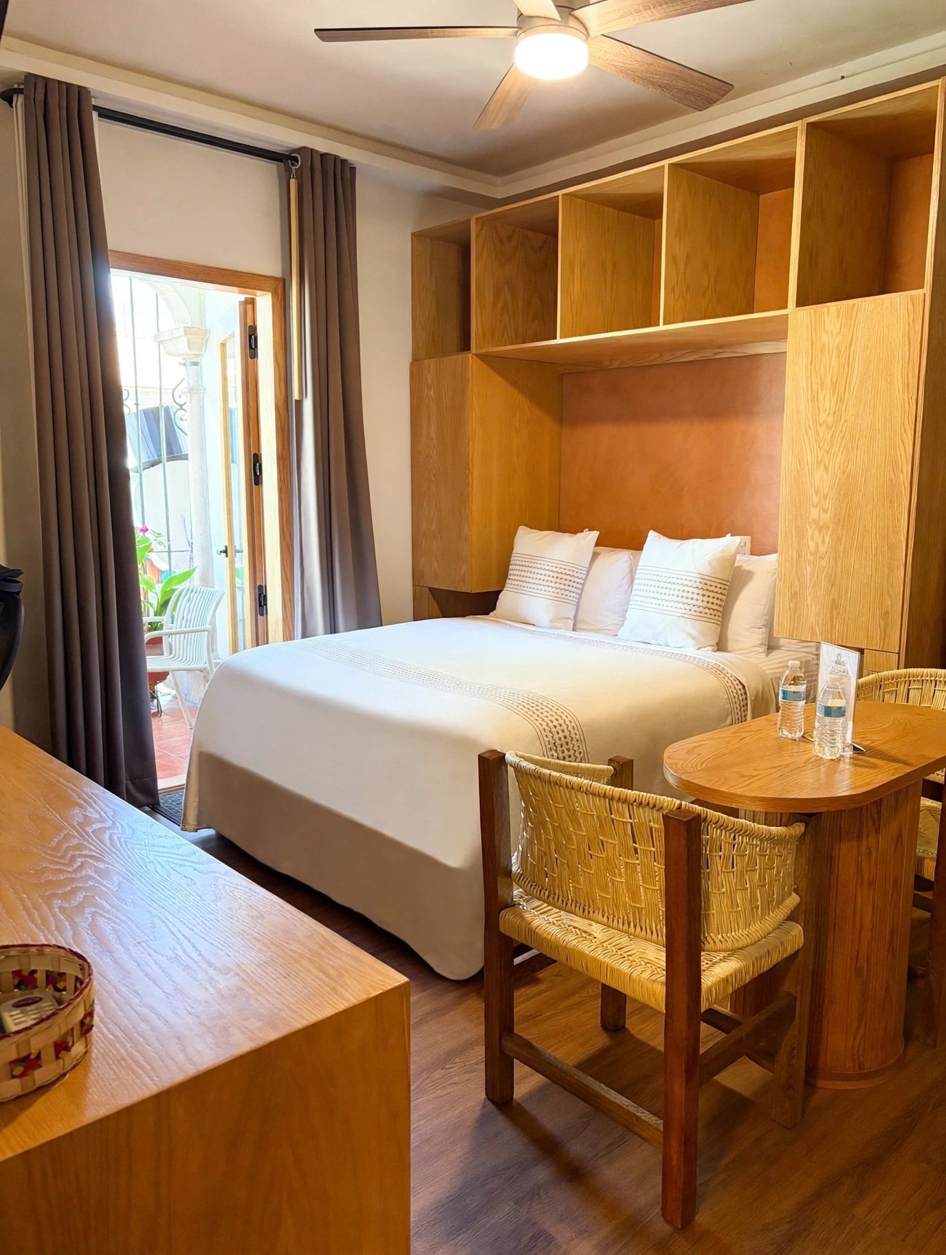 Une chambre d’hôtel avec un grand lit, une table à manger avec des chaises, un ventilateur de plafond et des étagères encastrées en bois.