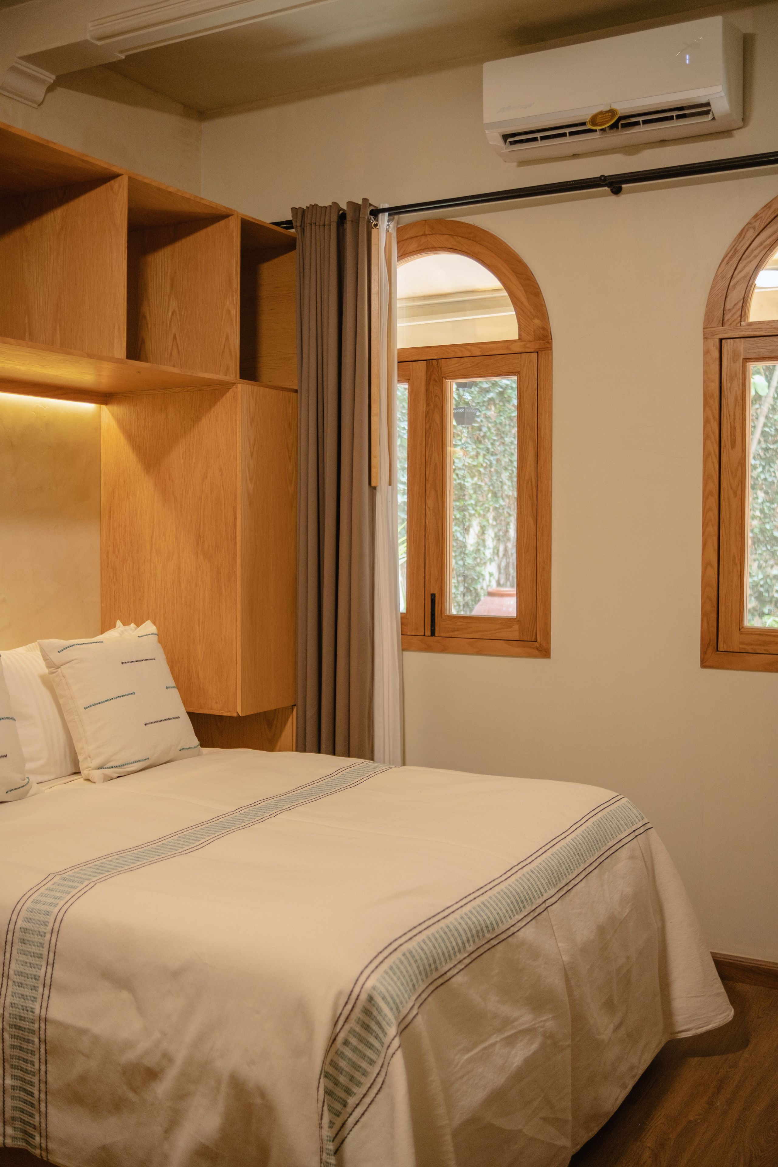 A bedroom with a bed, wooden headboard, two windows with wooden frames, and an air conditioning unit.