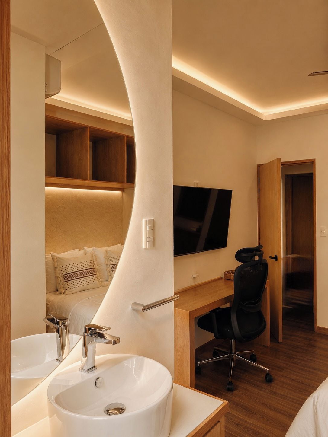 A modern hotel room with a curved wall, a large bed, a desk with a chair, a television, and a bathroom with a sink and mirror.