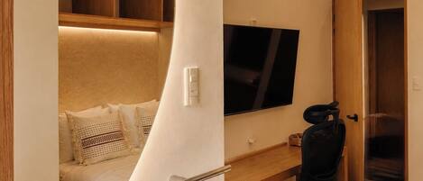A modern hotel room with a curved wall, a large bed, a desk with a chair, a television, and a bathroom with a sink and mirror.