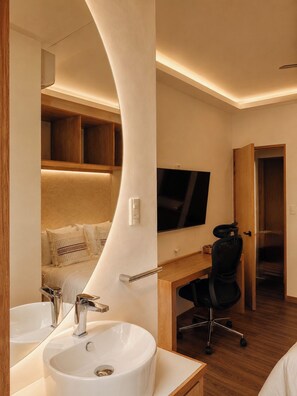 A modern hotel room with a curved wall, a large bed, a desk with a chair, a television, and a bathroom with a sink and mirror. - Don Manuel - Luxury Apartments & Studios (Oaxaca)