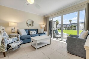 A living room with a blue sofa, a white coffee table, patterned armchairs, and a grey armchair. There is a large mirror on the wall, a ceiling fan, and a sliding glass door leading to an outdoor area with a lawn and a pool. - New Listing! Updated Oceanview 2/2 | Beachfront Pool & Beach Access (New Smyrna Beach)