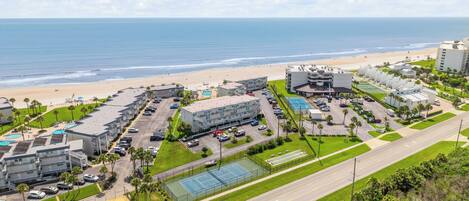 Vista aérea de un hotel costero con varias construcciones, una playa y una cancha de tenis.