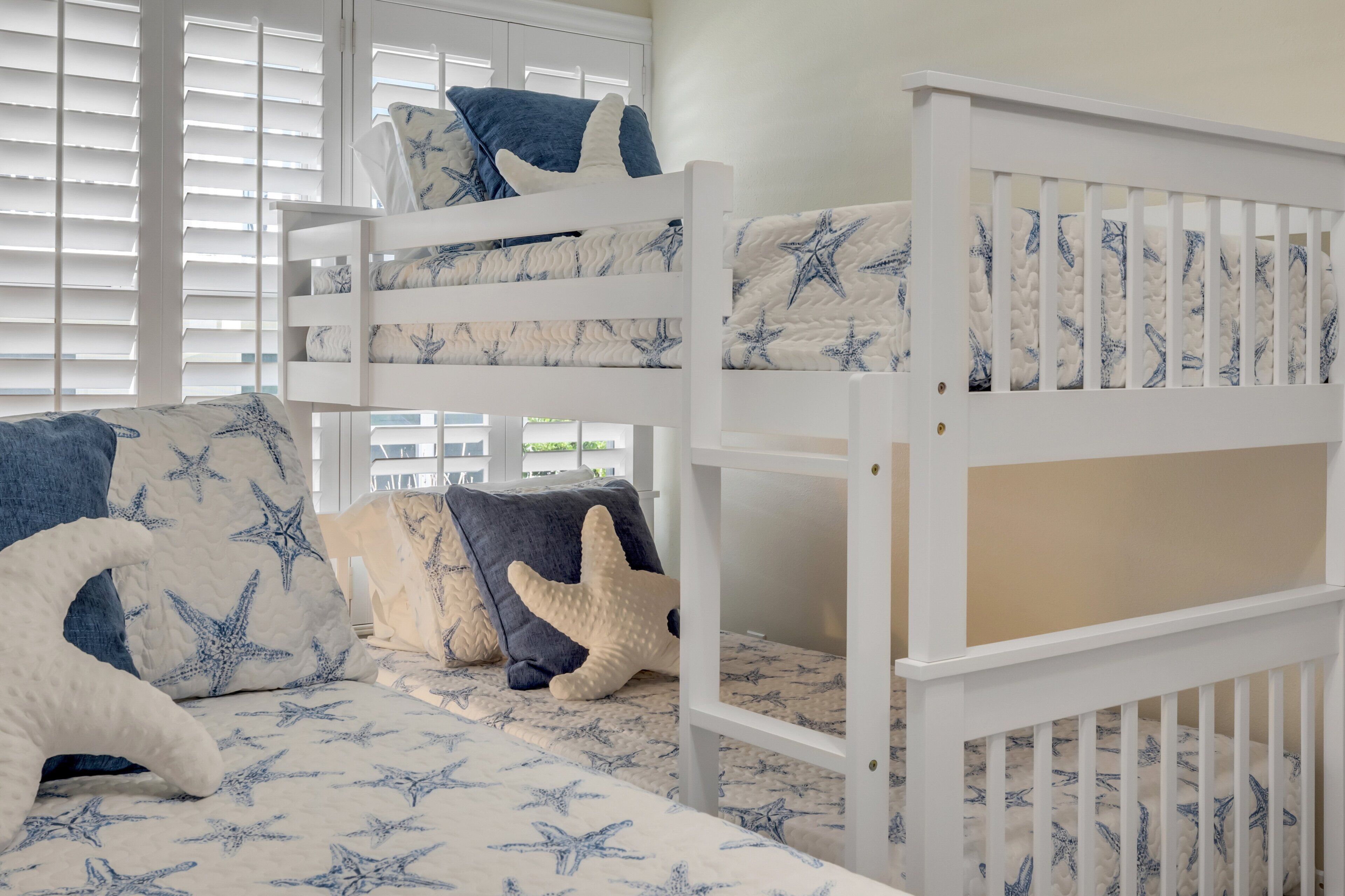 Uma cama beliche com colchão e almofadas estampados com tema de estrelas-do-mar, um berço branco e uma janela com persianas.