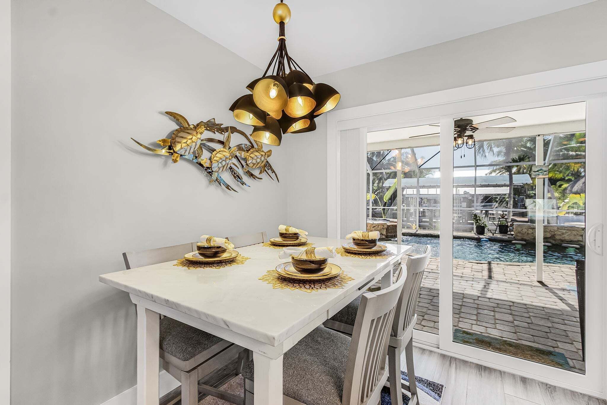 A dining area with a white table set for four, a decorative wall piece, and a view of a pool outside.