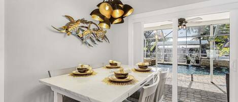 A dining area with a white table set for four, a decorative wall piece, and a view of a pool outside.