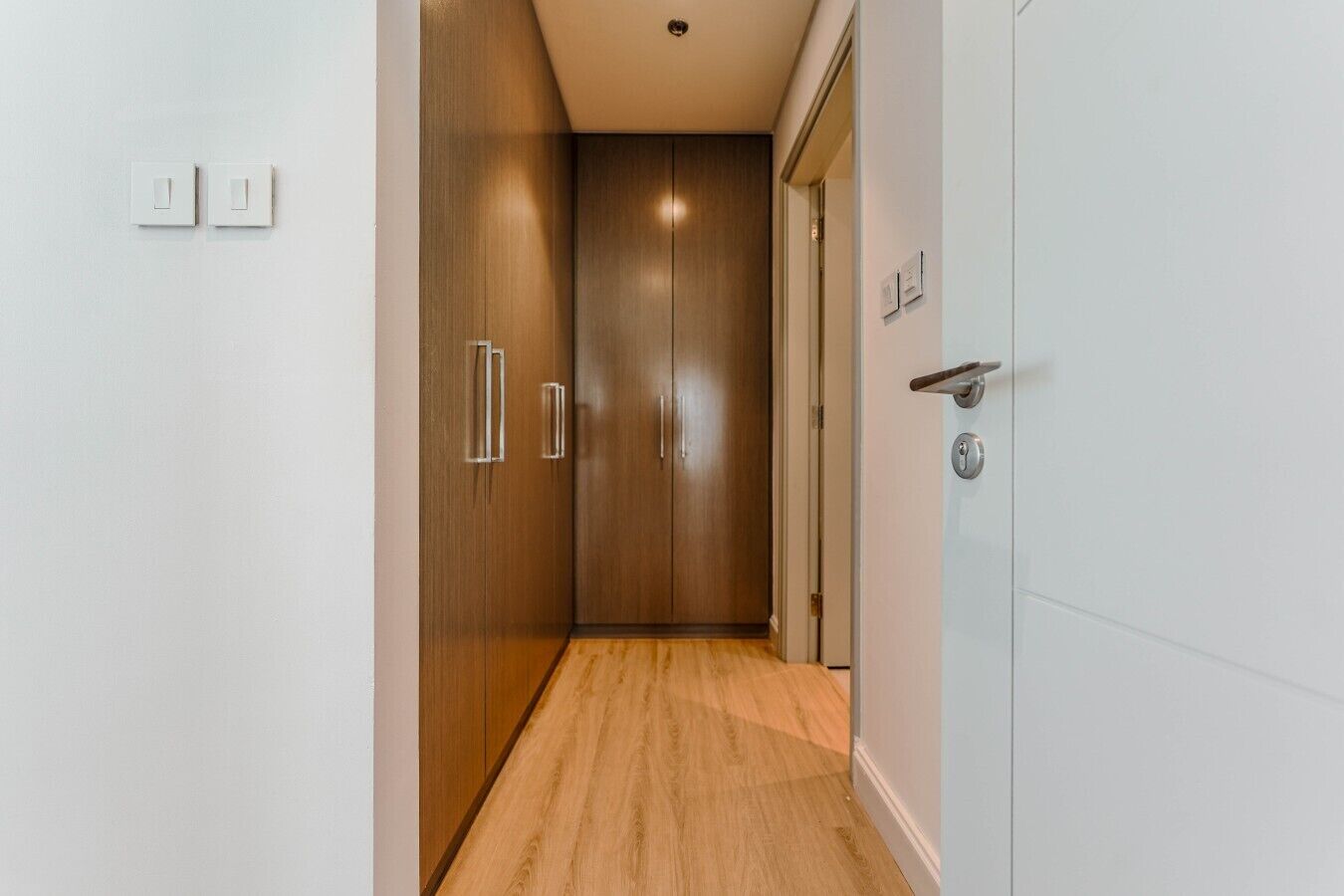 A hallway with wooden flooring, a door with a handle, and a light switch on the wall.