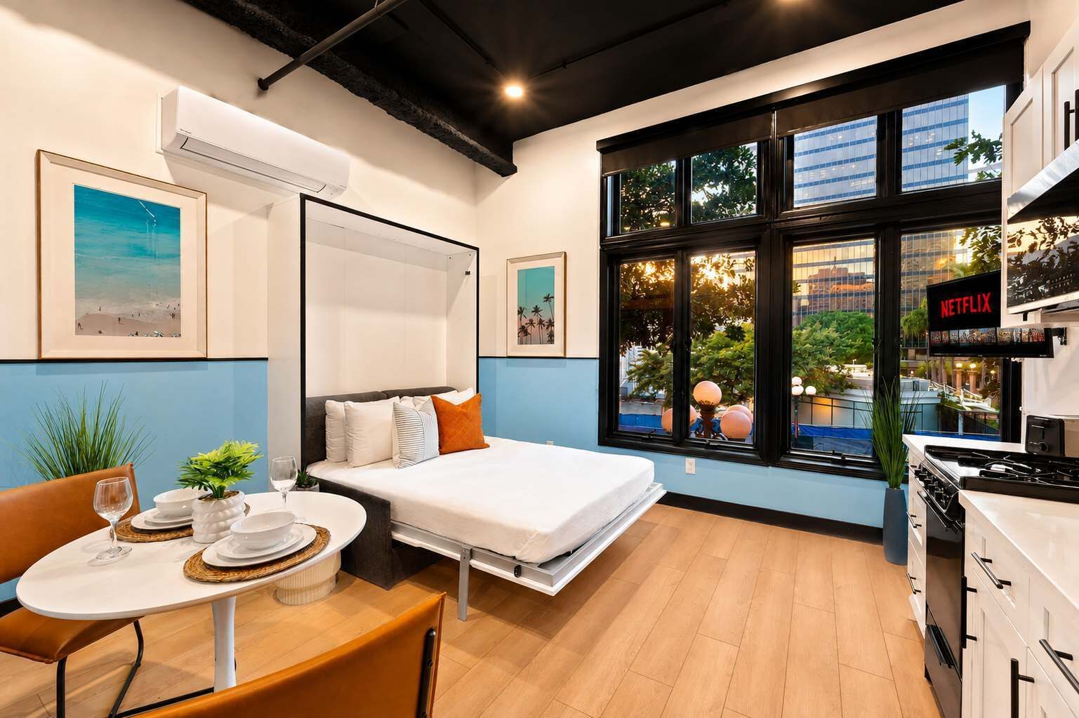 A modern hotel room with a bed, a dining table with chairs, a TV, and a window with a view of the city.