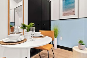 A dining area with a round table set for two, white plates, bowls, and a glass. There is a chair with a brown cushion, a potted plant, and a wall-mounted light fixture. - Petco Park Proximity | Urban Downtown Suite (San Diego)