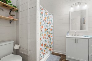 Baño con inodoro, lavabo y espejo. La cortina de ducha tiene un estampado de frutas.