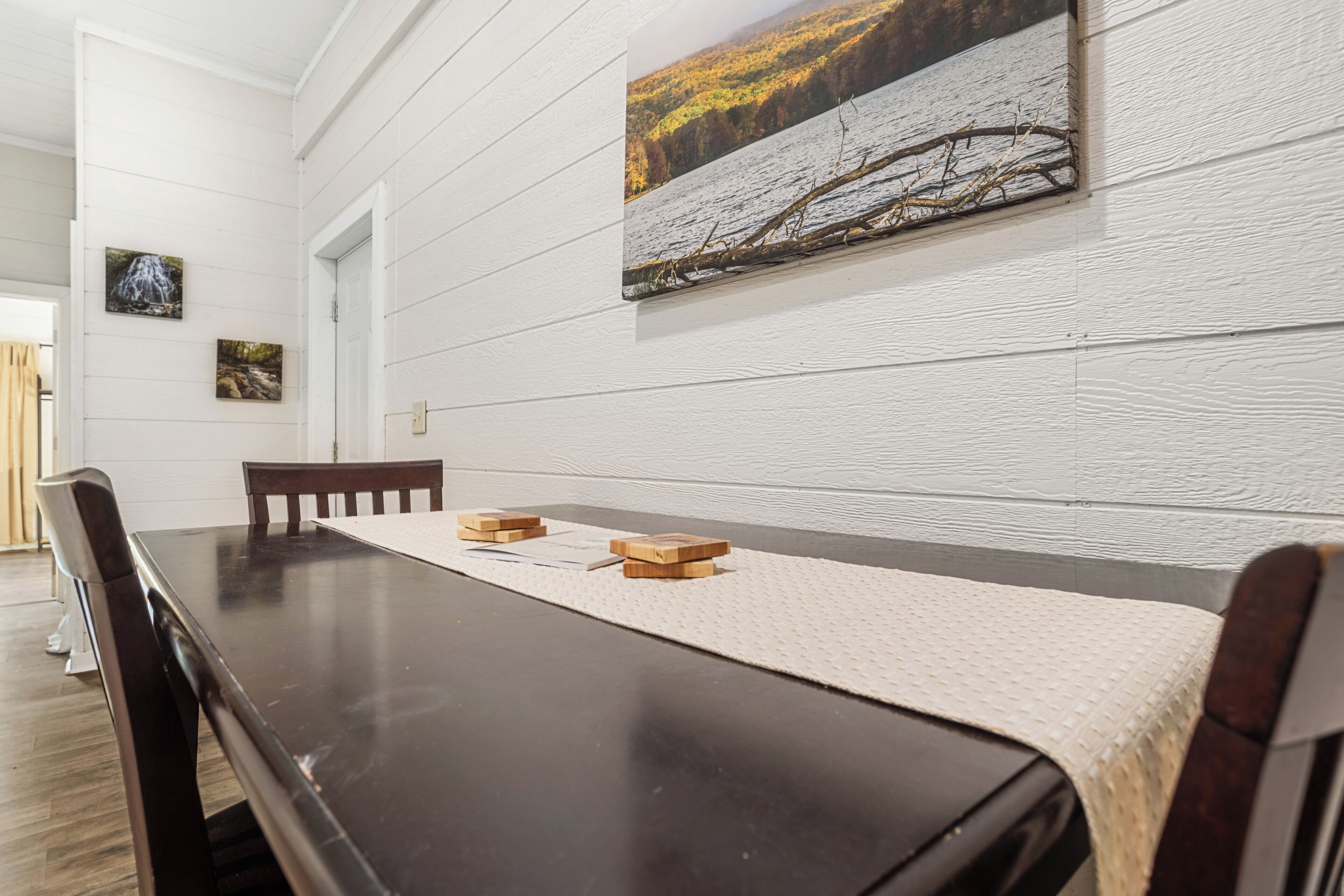 A dining room with a long table, wooden chairs, and a white textured table runner. There are two framed pictures on the wall above the table.
