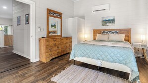 Ein Schlafzimmer mit einem hölzernen Schminktisch, einem Bett mit einer gesteppten blauen Bettdecke, einem Nachttisch mit einer Lampe und einer Wand-Klimaanlage.