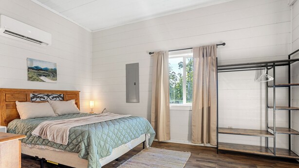 Ein Schlafzimmer mit Bett, Holzboden, einem Fenster mit Vorhängen, einer Wand-Klimaanlage und einem Regal.