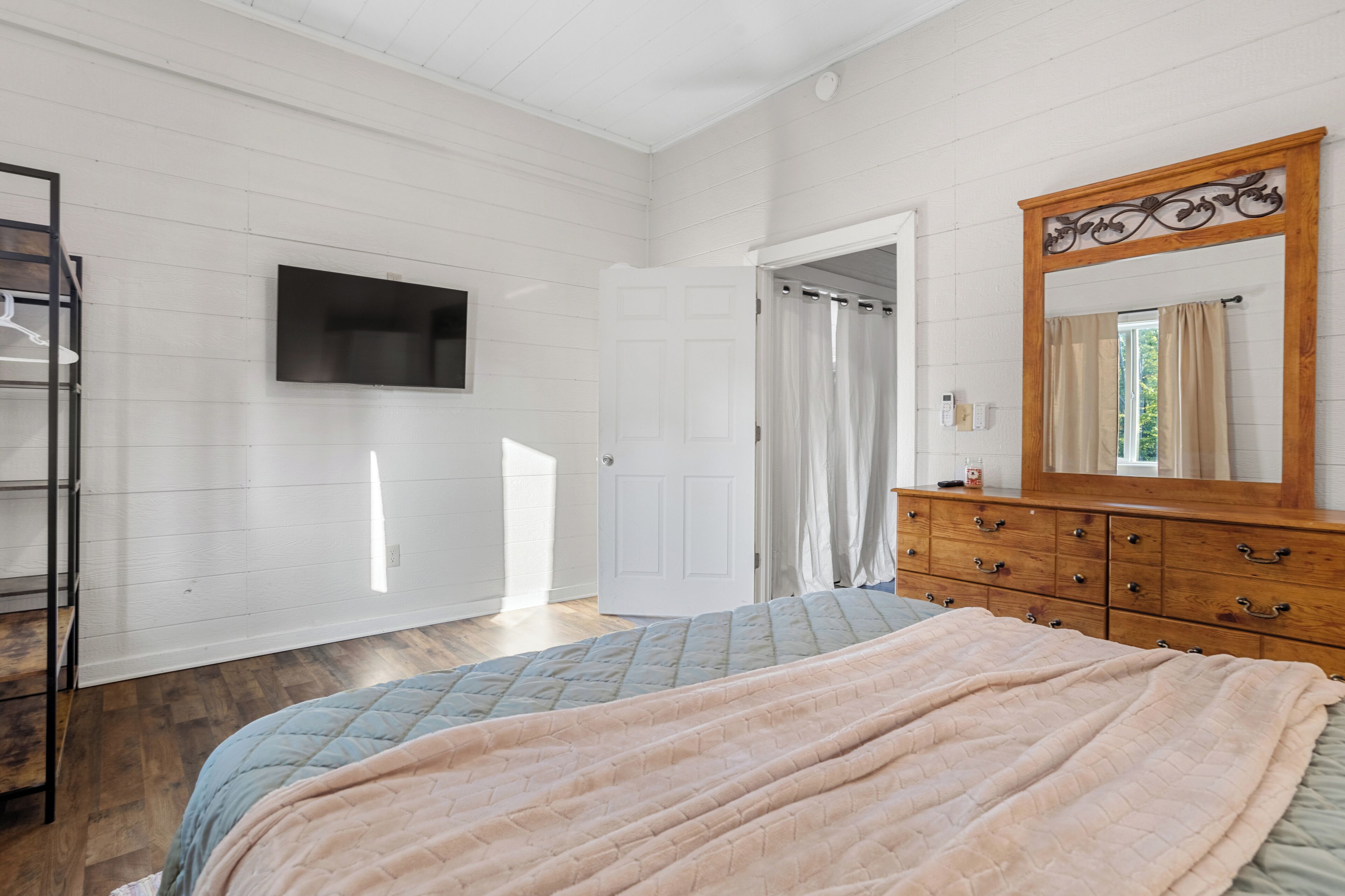 Un dormitorio con cama, cómoda, televisor montado en la pared y puerta que da a otra habitación.