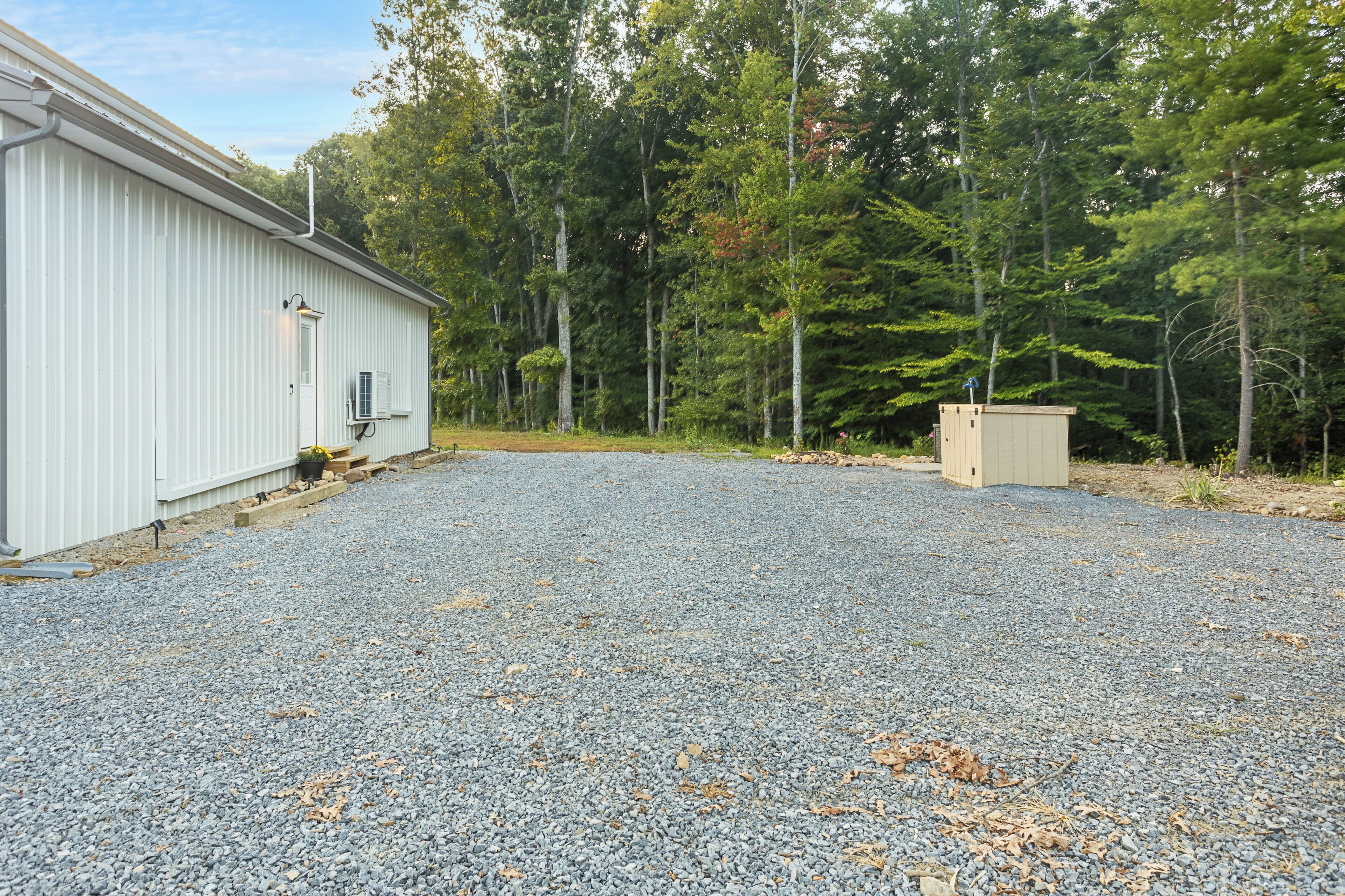 A gravel driveway leading to a building with a metal exterior and a small shed in a wooded area.