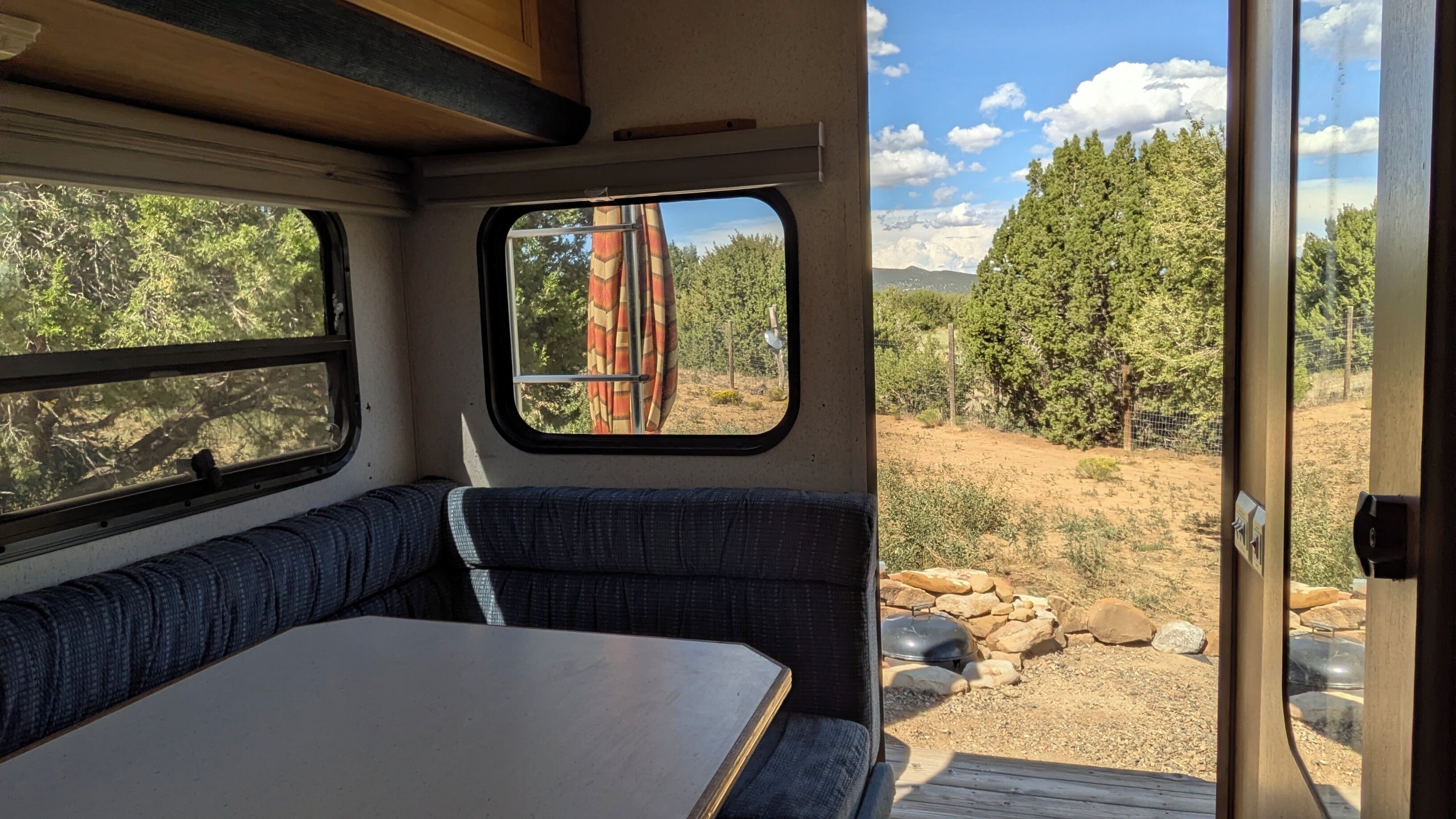 A campervan interior with a table and bench seating, a window with a view of a forest, and a door leading outside.