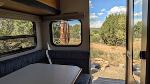 A campervan interior with a table and bench seating, a window with a view of a forest, and a door leading outside. - Mountain bungalow in serene Tijeras.  20 min. to downtown ABQ, 35 to Santa Fe (Tijeras)