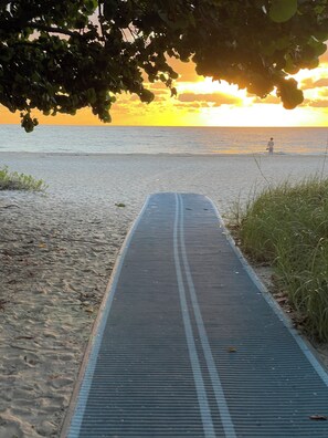 Ein Weg führt zu einem Strand, im Hintergrund ein Sonnenuntergang.
