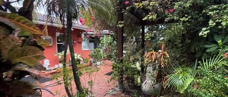 A brick pathway leading to a building with a red facade, surrounded by lush greenery and flowering plants.
