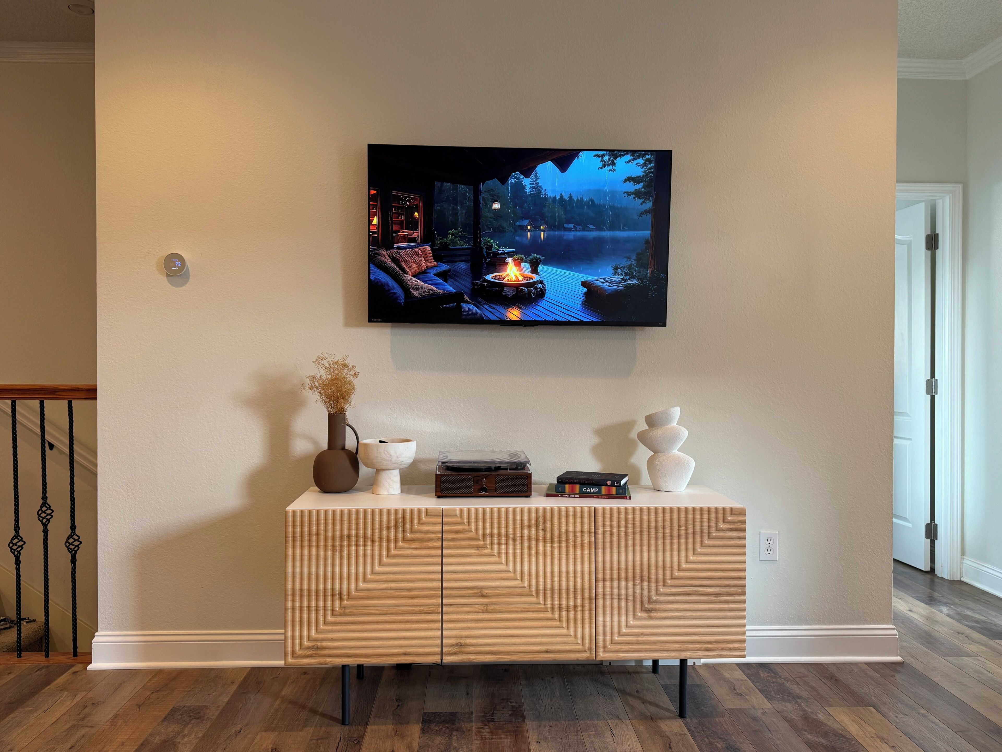 Een woonkamer met een flatscreen televisie aan de muur, een houten console met decoratieve objecten, en een trap met een leuning.