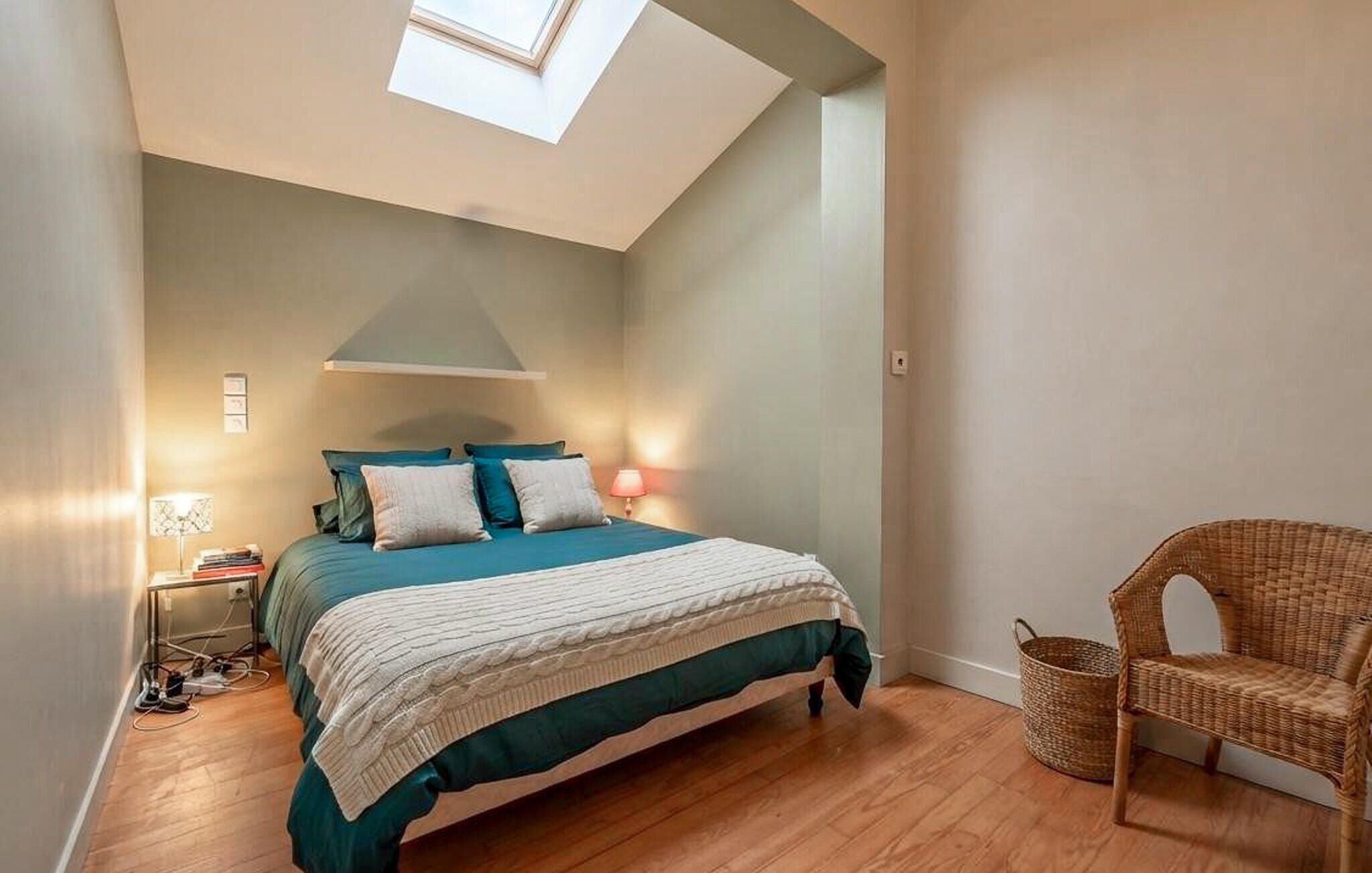 Ein Schlafzimmer mit Bett, Nachttischlampen, einem Korbstuhl und einem Dachfenster.