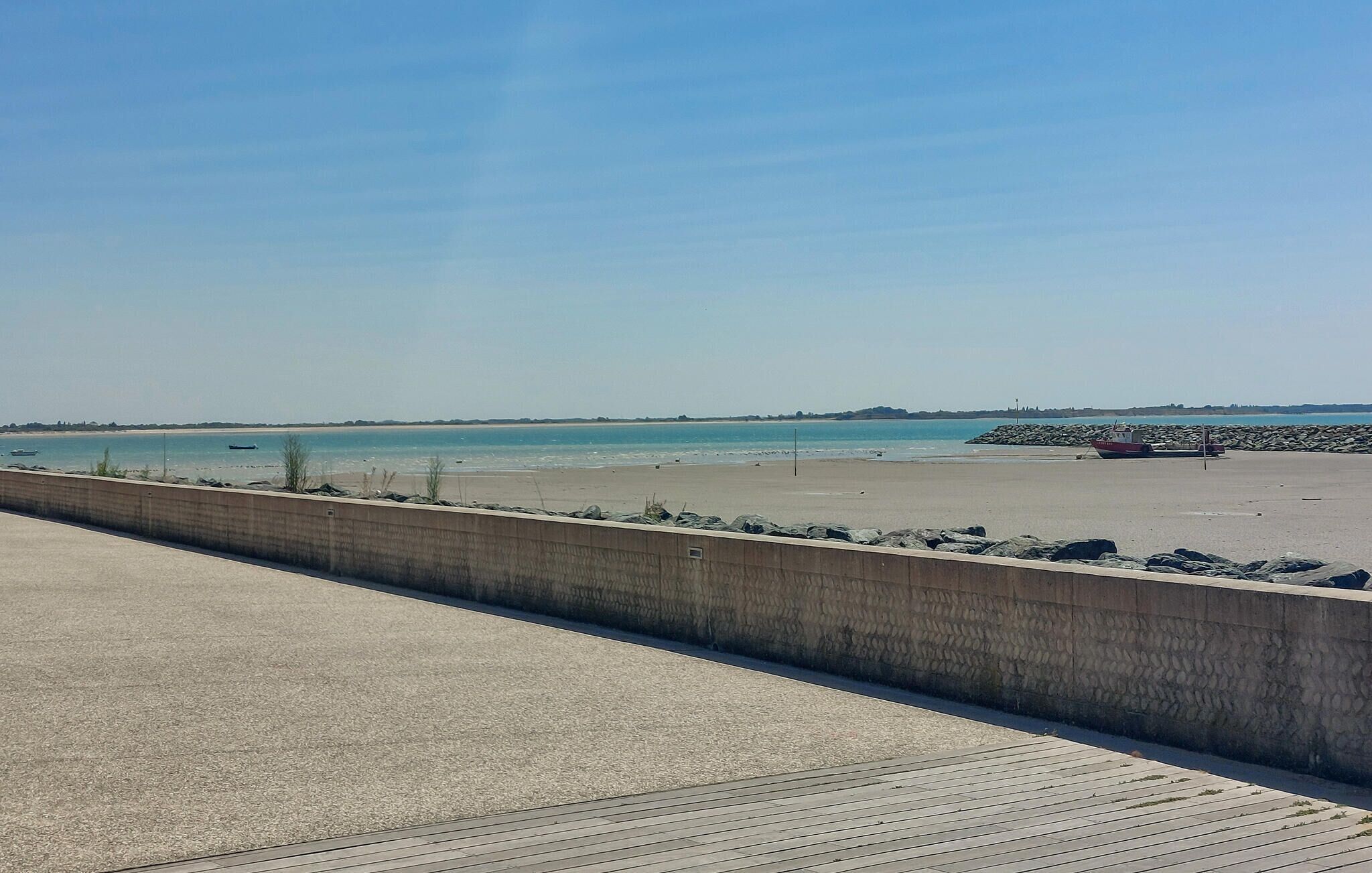 Een kustpad met een betonnen afscheiding, een zandstrand en een kalme zee.