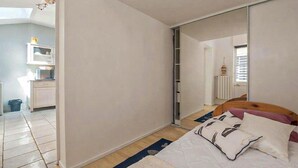 A bedroom with a bed, a wardrobe, a mirror, and a kitchenette in the background. - Charmante maison proche plage (Châtelaillon-Plage)