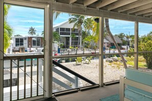 Vista desde el interior de un edificio con amplios ventanales que dan a una zona de piscina, un edificio con balcón y palmeras.