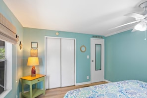 A bedroom with teal walls, a white door, a wooden nightstand with a lamp, a bed with a blue and white patterned bedspread, and a ceiling fan. - Waterfront Escape - Canal Front, 50ft Dock, King Beds and Pet Friendly (Little Torch Key)
