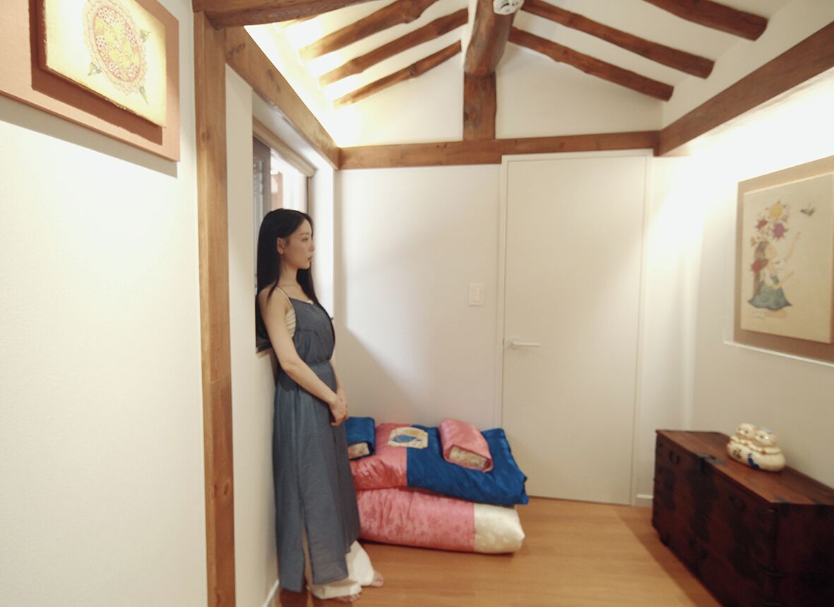 Une pièce avec un plafond en bois, un lit recouvert d’une couverture rose et bleue, et une commode en bois sur laquelle est posé un objet décoratif.