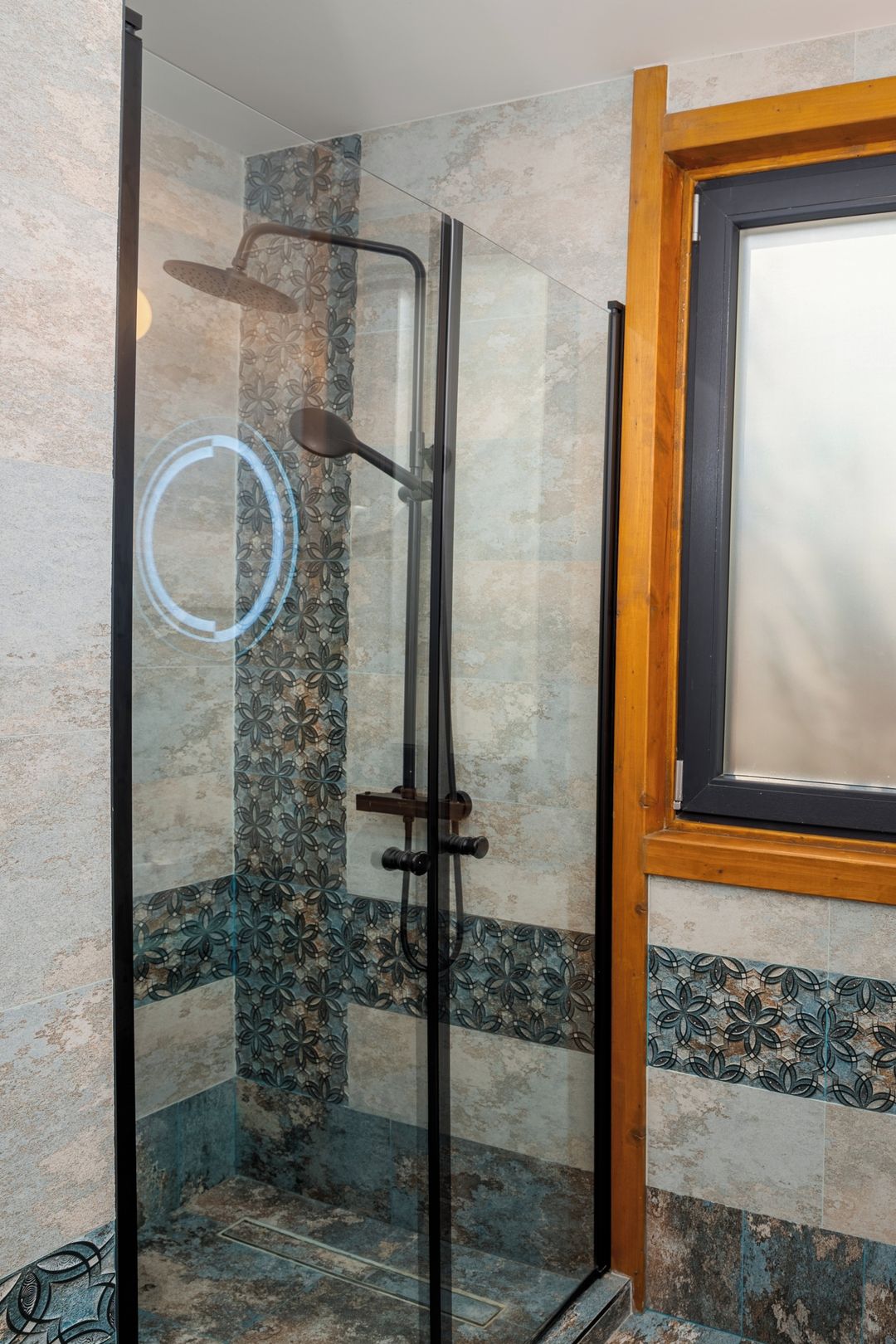 Een in glas omsloten douche met stortdouche, een houten deurkozijn en decoratieve tegels op de wanden.