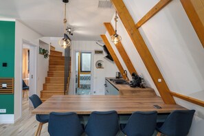 A modern kitchen with a wooden staircase, a dining area with a large table and chairs, and a view into the living space. - Vatra Verde - A-Frame Cabin (Trăisteni)